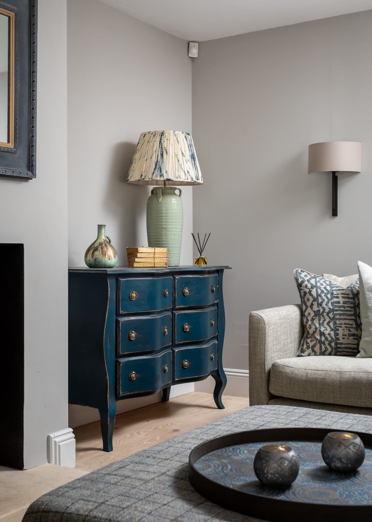 Antiqued blue painted chest in living room with corner sofa and footstool. Table lamp and silk Shibori shade