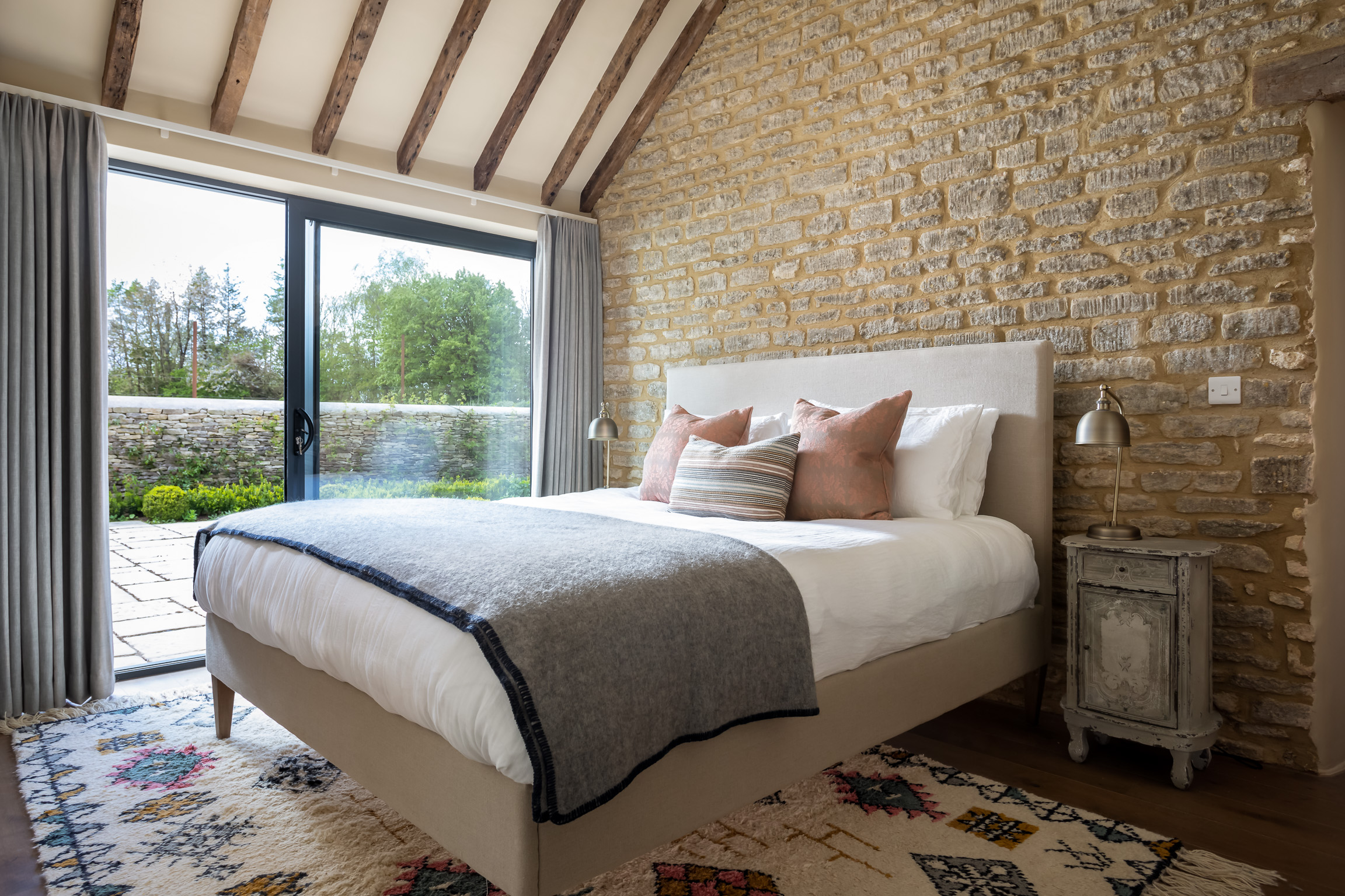 King size bed with fabric headboard, large contemporary Berber style rug. All against stone wall with glass sliding doors to garden.