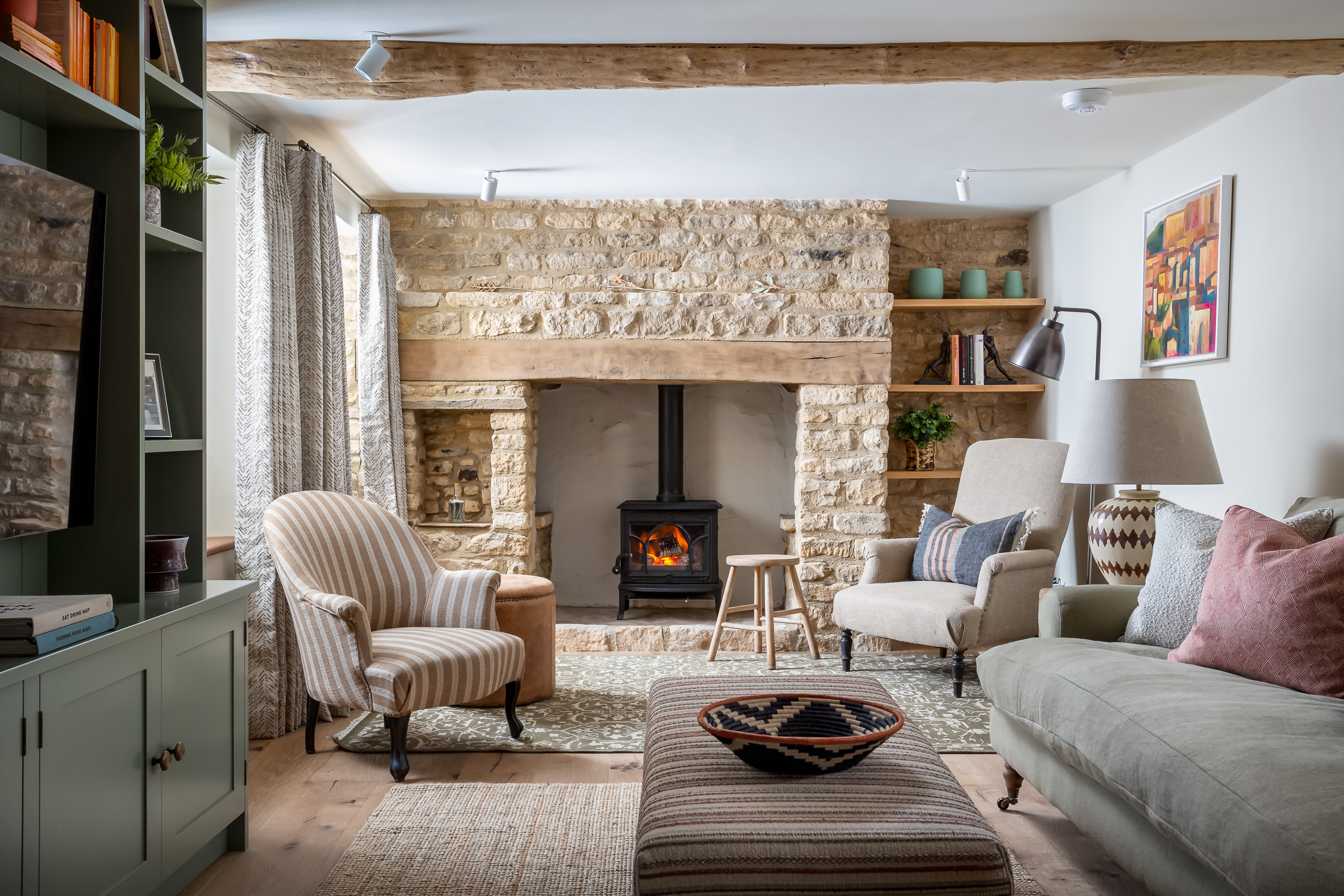 Living room with fireplace and stone feather wall. Hand made curtains, bespoke striped footstool, linen upholstered armchairs. Green patterned wool rug