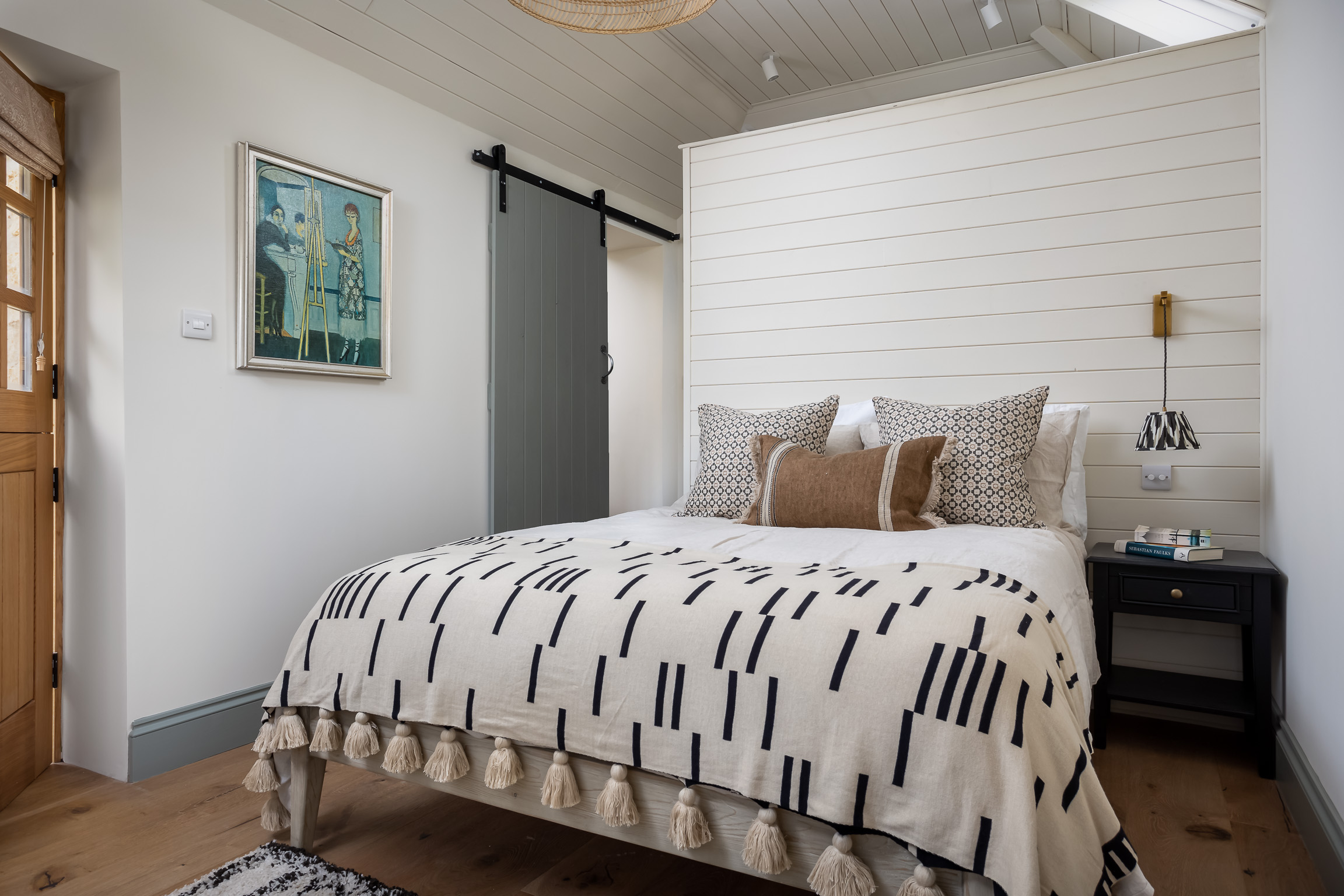 Annexe bedroom with barn door to bathroom; divider wall behind bed; black & cream bed throw, cushions on bed in strong earth tones