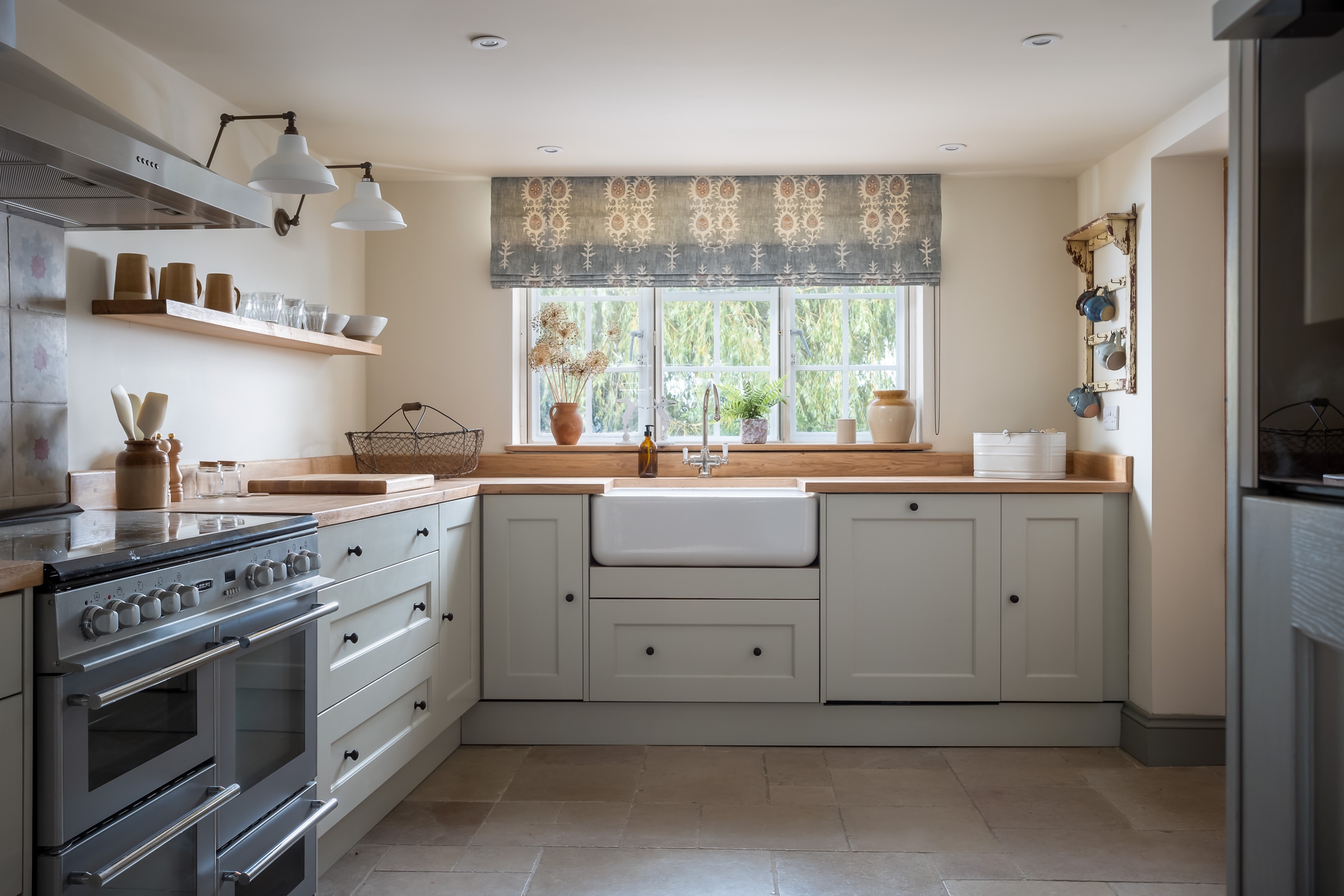 Limestone colour kitchen with oak work surface. Stone floor, Belfast sink and roman blinds at windows.