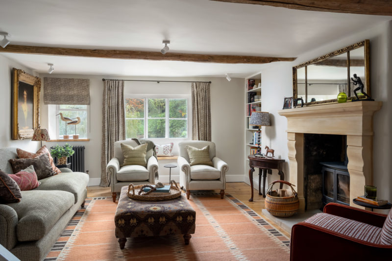 sitting room with large footstool on terracotta rug, pair comfy armchairs, stone fireplace, D end table & lamp, bookshelves, wing back chair in burnt orange