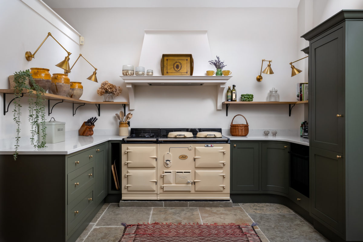 Farmhouse kitchen with green units, quartz surface, brass wall lights, cream Aga, stone floor and antique rug