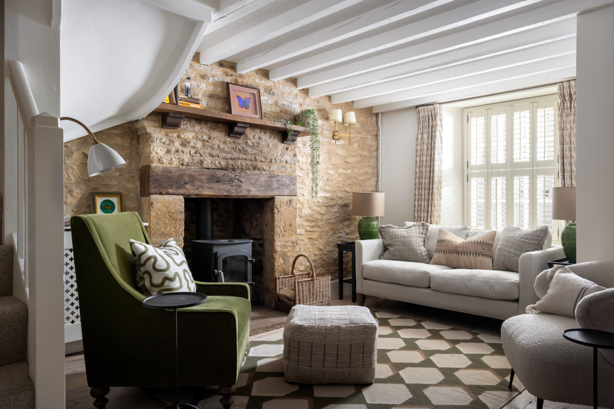 Stone walls. pine floorboards, neutral sofa with earthy colour cushions, green pattern rug, olive velvet armchair, footstool, boucle tub chair. Curtains at front window in green & ochre