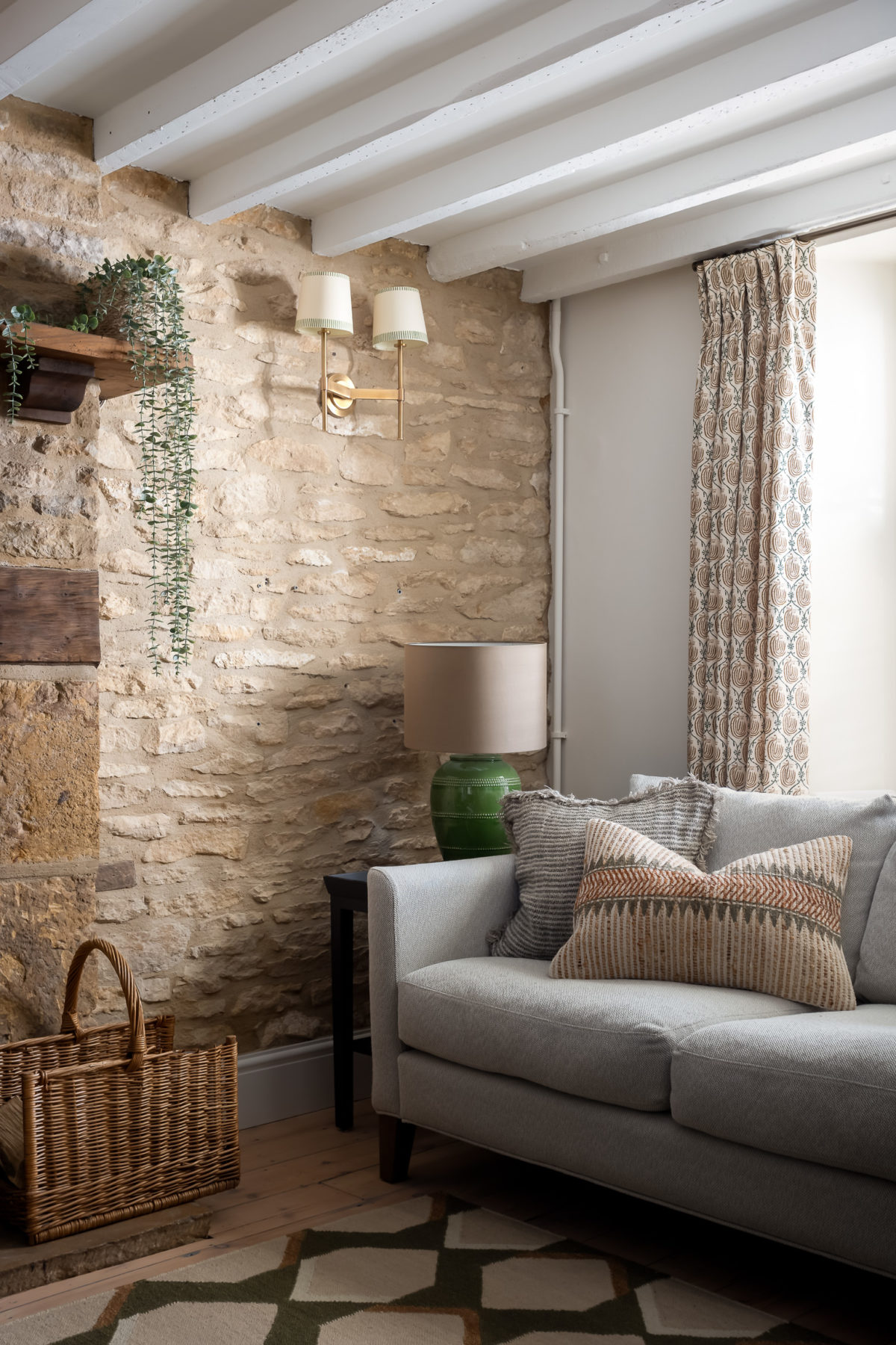 Grey sofa with green, cream & orange cushions, stone wall, brass wall lights with cream card shades, open fireplace & log basket