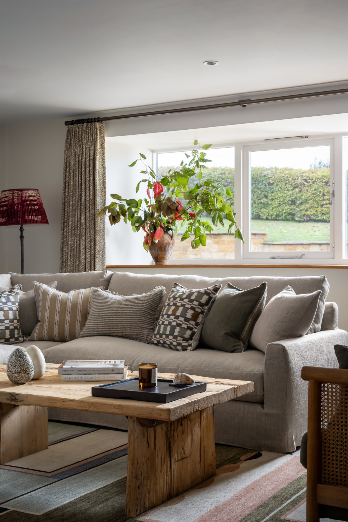 Living room in barn, oak coffee table, neutral corner sofa with mis of green, terracotta & taupe cushions. Floor lamp.